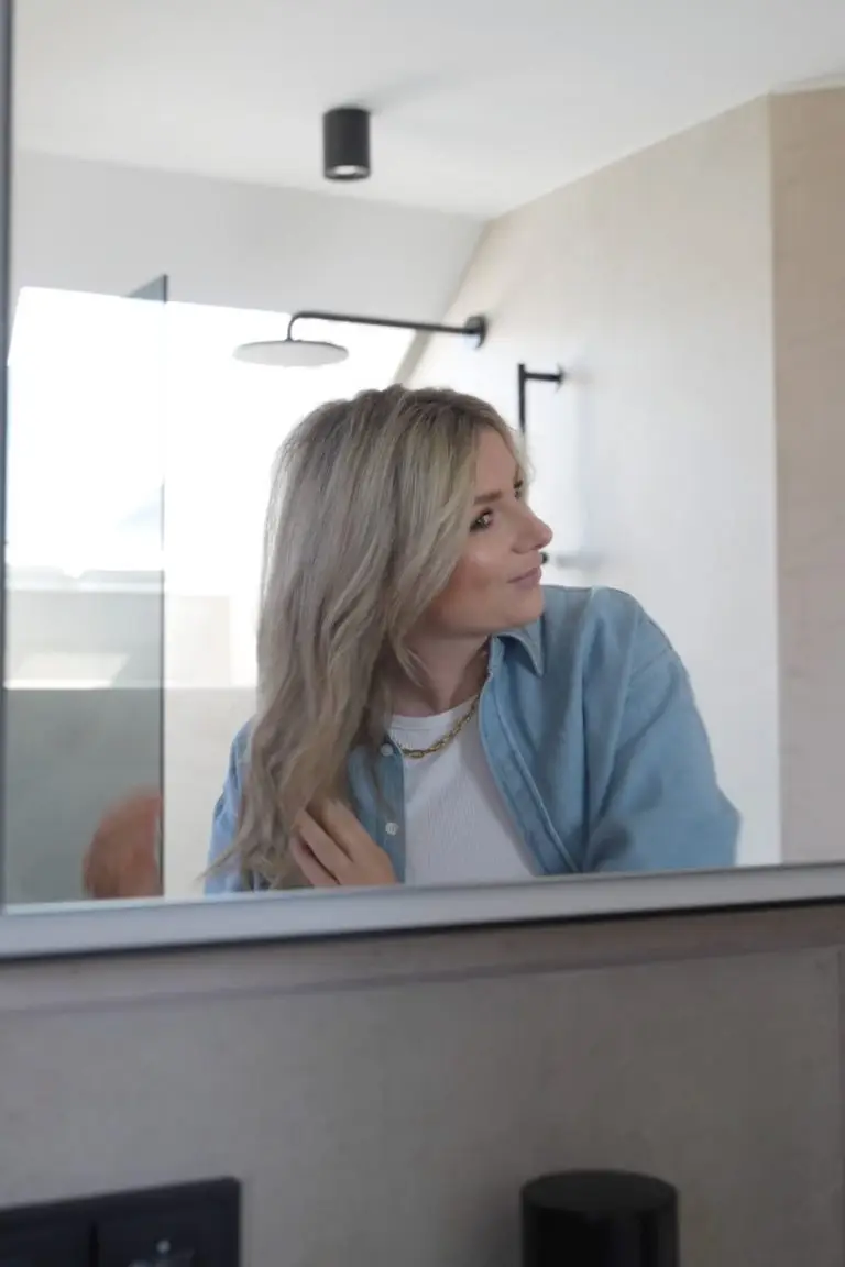 Blonde Frau betrachtet sich im Spiegel und kämmt sich durch die Haare